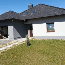 Dwukondygnacyjny dom z szarą i białą elewacją, ciemną dachówką, dwoma oknami dachowymi, brązowymi ramami okiennymi i drzwiami wejściowymi. Widoczny trawnik, ścieżka z płyt i otwarty garaż.