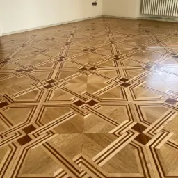 Wzór parkietu z jasnego drewna z ciemniejszymi, geometrycznymi wstawkami w pustym pokoju z grzejnikiem i gniazdkiem elektrycznym.