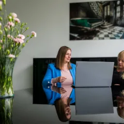 Dwie kobiety przy laptopie na tle biura, kwiaty w wazonie. Refleksja w blacie stołu. Konsultacja biznesowa, analiza danych lub spotkanie zespołu.