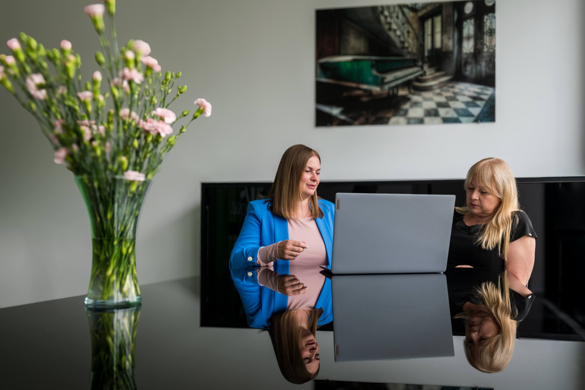 Dwie kobiety przy laptopie na tle biura, kwiaty w wazonie. Refleksja w blacie stołu. Konsultacja biznesowa, analiza danych lub spotkanie zespołu.