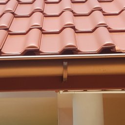 DGS - Fragment dachu z ceramicznej dachówki w odcieniach czerwieni i brązu, z widoczną rynną w kolorze miedzianym, zamocowaną do okapu.