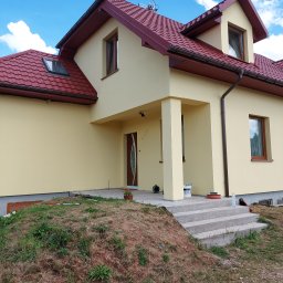 DGS - Świeżo otynkowany dom jednorodzinny z czerwonym dachem i brązowymi rynnami, widoczne schody wejściowe i pozostałości po pracach wykończeniowych.