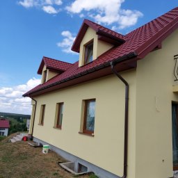 DGS - Żółta elewacja domu jednorodzinnego z brązowymi oknami, dachem z czerwonej dachówki i brązowymi rynnami w słoneczny dzień. Widoczny fragment podwórka z wiadrem po farbie.