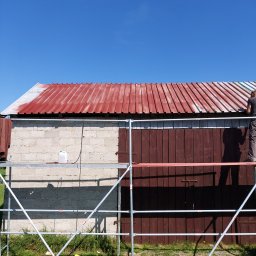 DGS - Mężczyzna maluje metalowy dach budynku na czerwono, stojąc na rusztowaniu; widoczny fragment ściany z pustaków i drewniane drzwi.