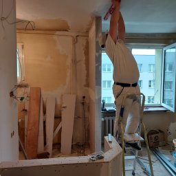 DGS - Pracownik w białym kombinezonie na aluminiowej drabinie, przykłada poziomicę do sufitu w trakcie remontu mieszkania, widoczne odsłonięte kable, płyty gipsowo-kartonowe i narzędzia.