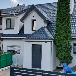 Nowoczesny dom jednorodzinny z szarą stolarką okienną i drzwiową, świeżo pomalowaną elewacją w odcieniach bieli, widoczny zza grafitowego ogrodzenia panelowego.