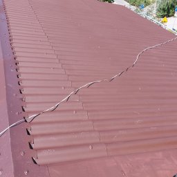 DGS - Widok z góry na nowo pomalowany, brązowy dach z blachy falistej, z widocznymi śladami farby na powierzchni, biegnący po dachu biały kabel i fragment cienia osoby w prawym dolnym rogu.