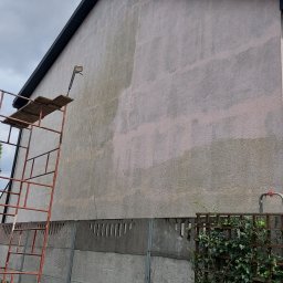 DGS - Fragment elewacji domu z widocznym efektem częściowego umycia pod ciśnieniem, z lewej strony rusztowanie z drewnianą platformą, w tle pochmurne niebo.