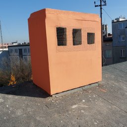 DGS - Pomarańczowa nadbudowa na dachu budynku w Kielcach, z trzema małymi oknami zabezpieczonymi siatką, antena na szczycie, błękitne niebo w tle.