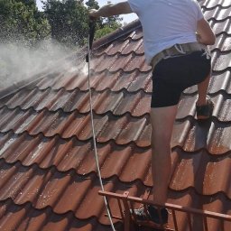 DGS - Pracownik w białej koszulce i czarnych spodenkach, zabezpieczony pasem, czyści czerwoną dachówkę myjką ciśnieniową, stojąc na małej drabince dachowej; w tle widać zielone drzewa i błękitne niebo.