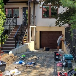 DGS - Mężczyzna maluje czarny metalowy płot przy domu z brązową bramą garażową, na brukowanym podjeździe leżą narzędzia i akcesoria malarskie.