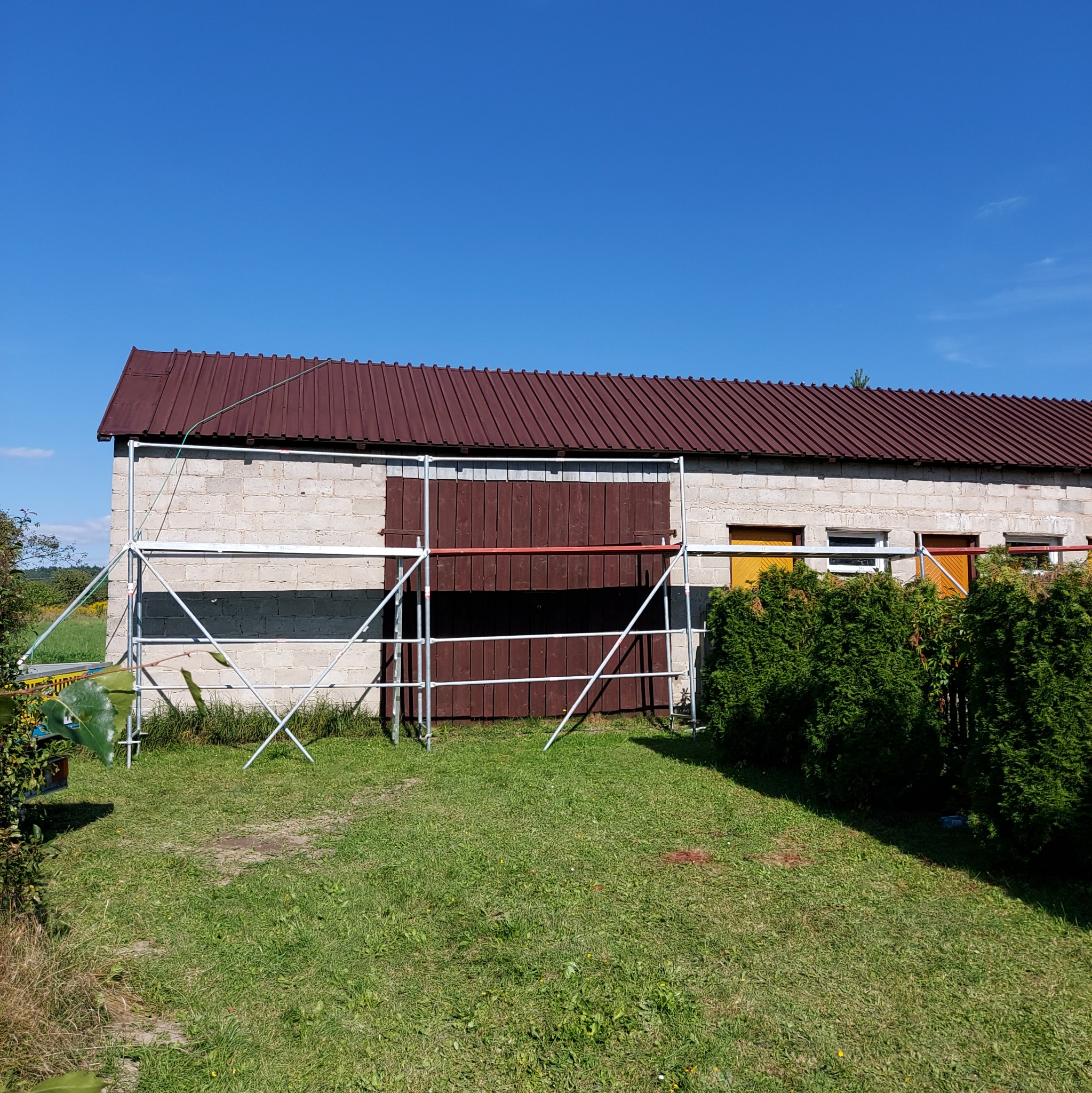 Budynek gospodarczy z szarej cegły z brązowym dachem i brązowymi drzwiami, częściowo zasłonięty rusztowaniem na tle zielonego trawnika i błękitnego nieba.