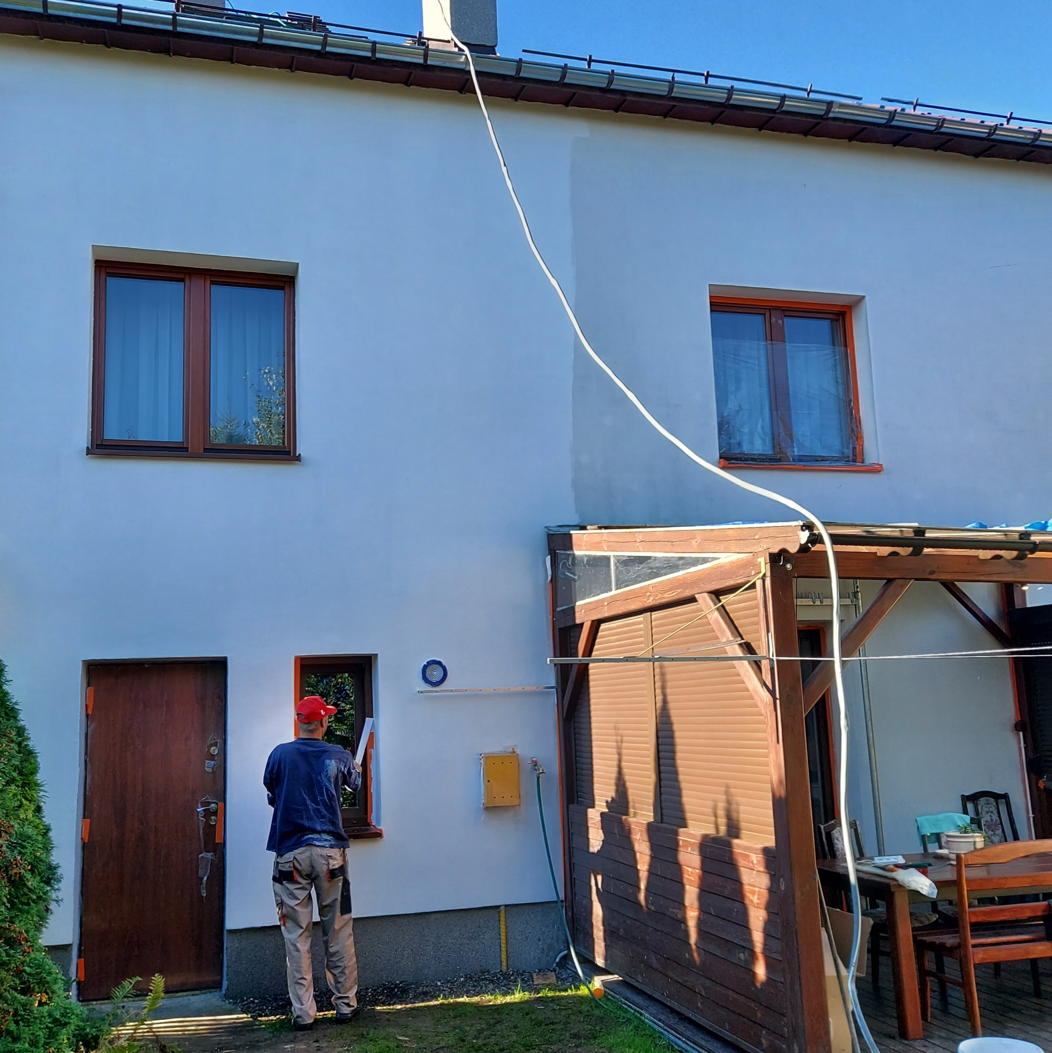 Pracownik w czerwonej czapce przygotowuje okno do malowania na białej elewacji domu, widoczna zakryta folią szyba, kabel od urządzenia oraz zielony wąż ogrodowy.
