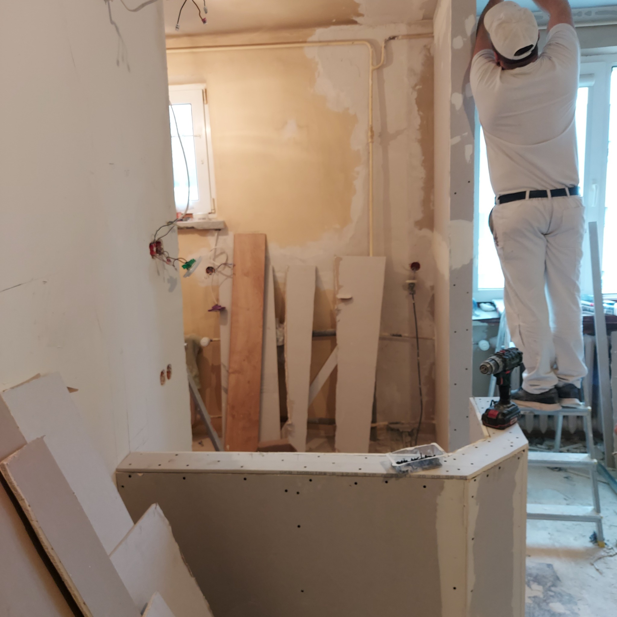 Pracownik w białym kombinezonie na drabinie montuje płytę gipsowo-kartonową na ścianie w trakcie remontu, widoczne kable elektryczne, płyty GK na podłodze i narzędzia.