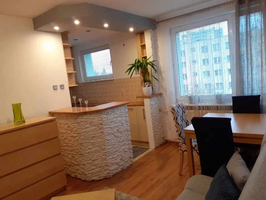 Widok na wnętrze mieszkania z aneksem kuchennym, barek z wykończeniem imitującym kamień, drewniany stół z krzesłami, okno z widokiem na budynek za oknem.