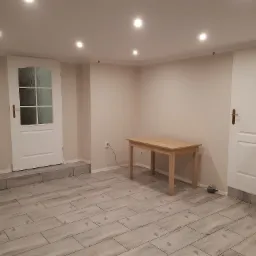 Jasne wnętrze po remoncie z nową podłogą z płytek imitujących drewno, drewnianym stołem, białymi drzwiami i oświetleniem punktowym w suficie.