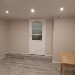 Wykończone wnętrze z panelami podłogowymi imitującymi drewno, białą stolarką drzwiową i jasnymi ścianami, oświetlone punktowo, z drewnianym stolikiem w rogu.