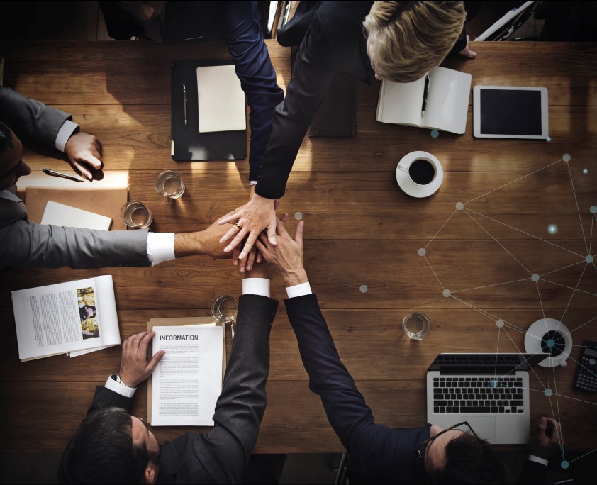 Widok z góry na drewniany stół konferencyjny z dłońmi kilku osób w garniturach, złożonymi na sobie w geście współpracy; obok laptop, tablet, notatniki, szklanki z wodą i filiżanka kawy.