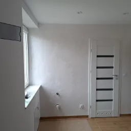 Świeżo wykończone wnętrze z gładzią w odcieniach bieli, z zamontowanymi drzwiami wewnętrznymi z poziomymi przeszkleniami, widoczne przyłącza wodne i gniazdko elektryczne, podłoga wyłożona płytkami...