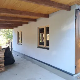 Wykończona elewacja budynku z drewnianymi oknami i przesuwną bramą garażową, widoczny stos drewna i czarny worek na zewnątrz.