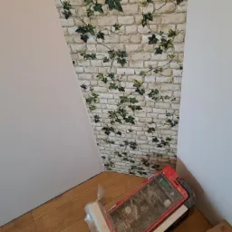 Fragment pokoju po położeniu tapety w cegiełki z motywem bluszczu na skośnej ścianie, widoczna skrzynka z narzędziami na podłodze.