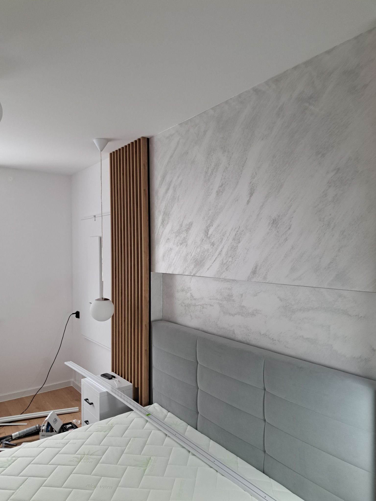 Ściana w sypialni z dekoracyjną strukturą imitującą marmur, drewniana ażurowa ścianka działowa i szary tapicerowany zagłówek łóżka, w trakcie montażu listew wykończeniowych.