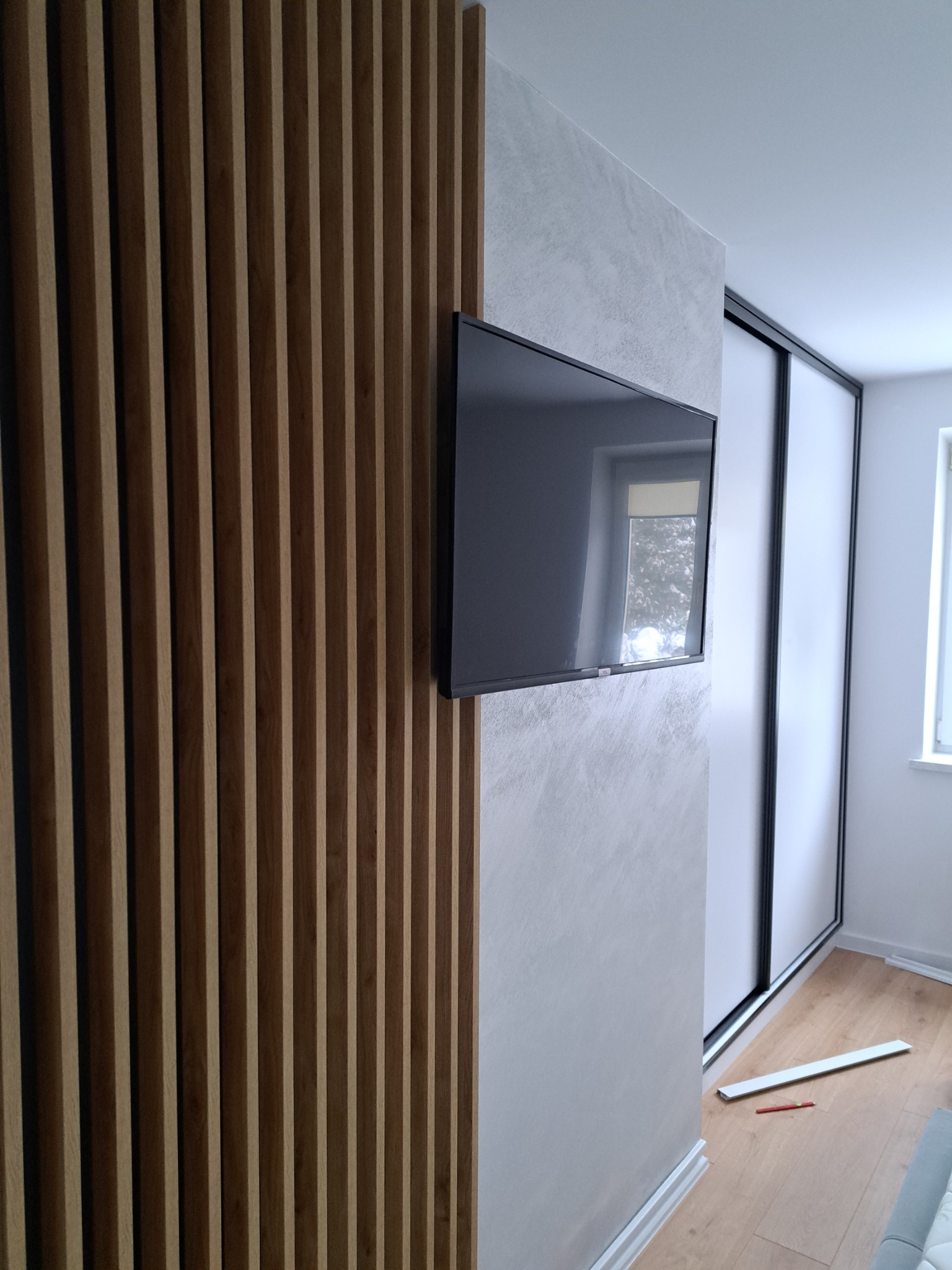 Telewizor zamontowany na ścianie z efektem betonu architektonicznego, obok drewniana ażurowa ścianka działowa i szafa przesuwna w Przysiekach.