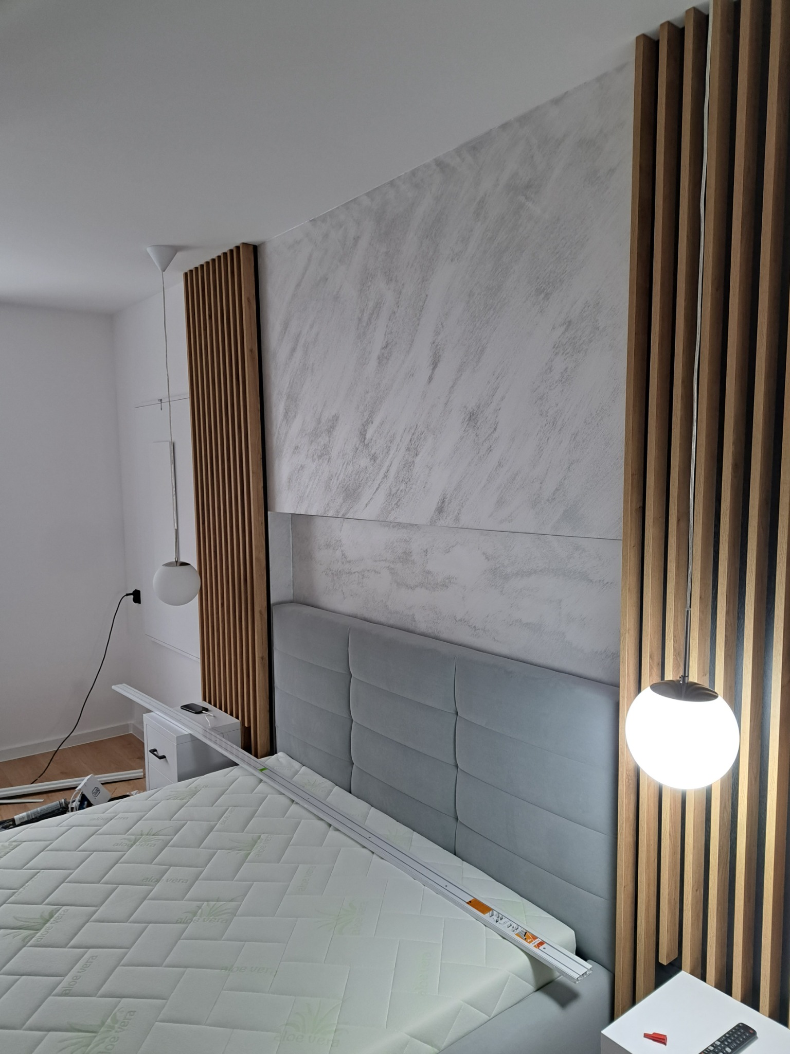 Ściana za łóżkiem z dekoracyjną strukturą imitującą marmur, panele drewniane pionowe po bokach, lampy wiszące z kulistymi kloszami, łóżko z szarym zagłówkiem i materacem w pokrowcu z nadrukiem Aloe...