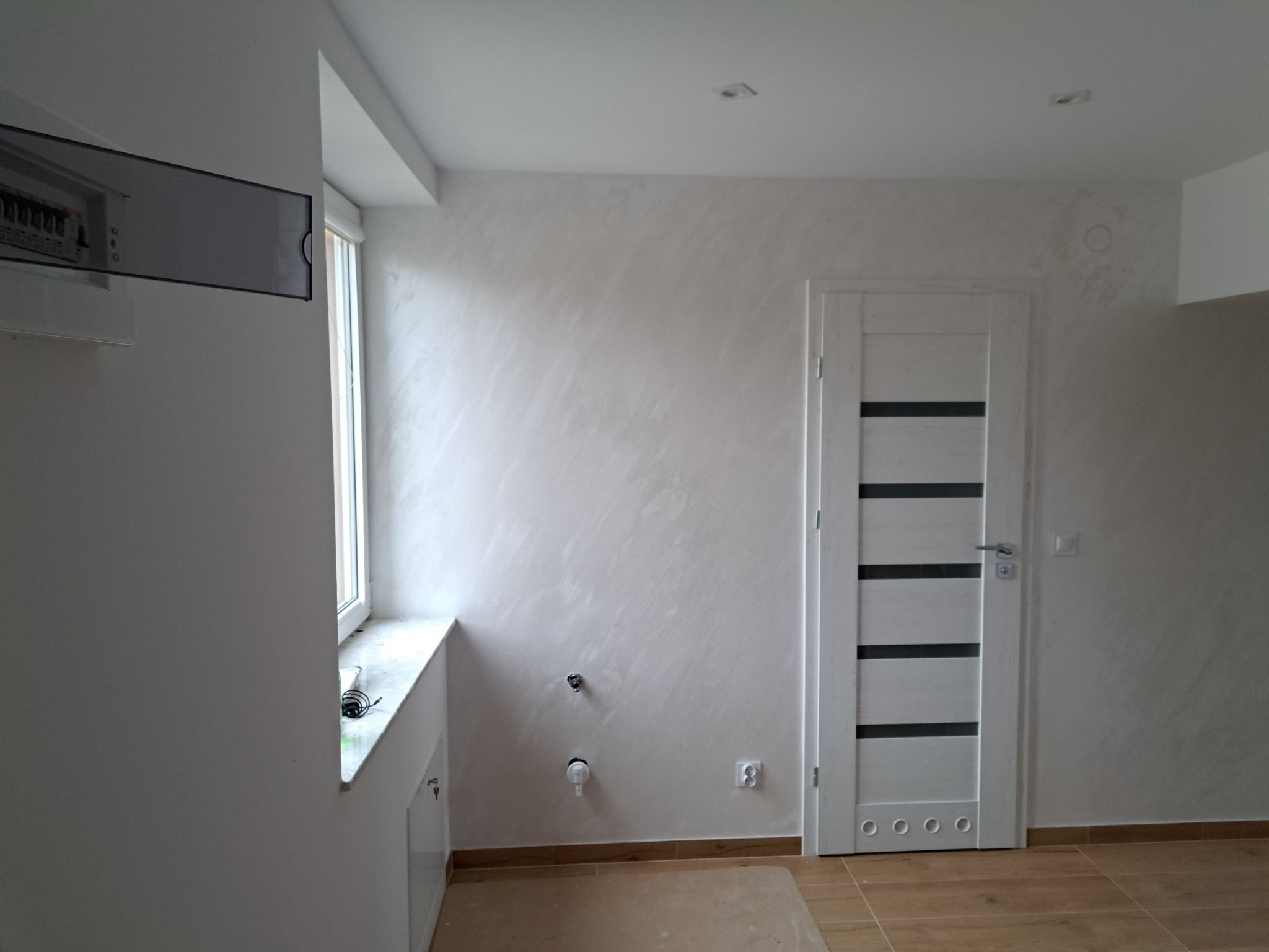 Świeżo wykończone wnętrze z gładzią w odcieniach bieli, z zamontowanymi drzwiami wewnętrznymi z poziomymi przeszkleniami, widoczne przyłącza wodne i gniazdko elektryczne, podłoga wyłożona płytkami...