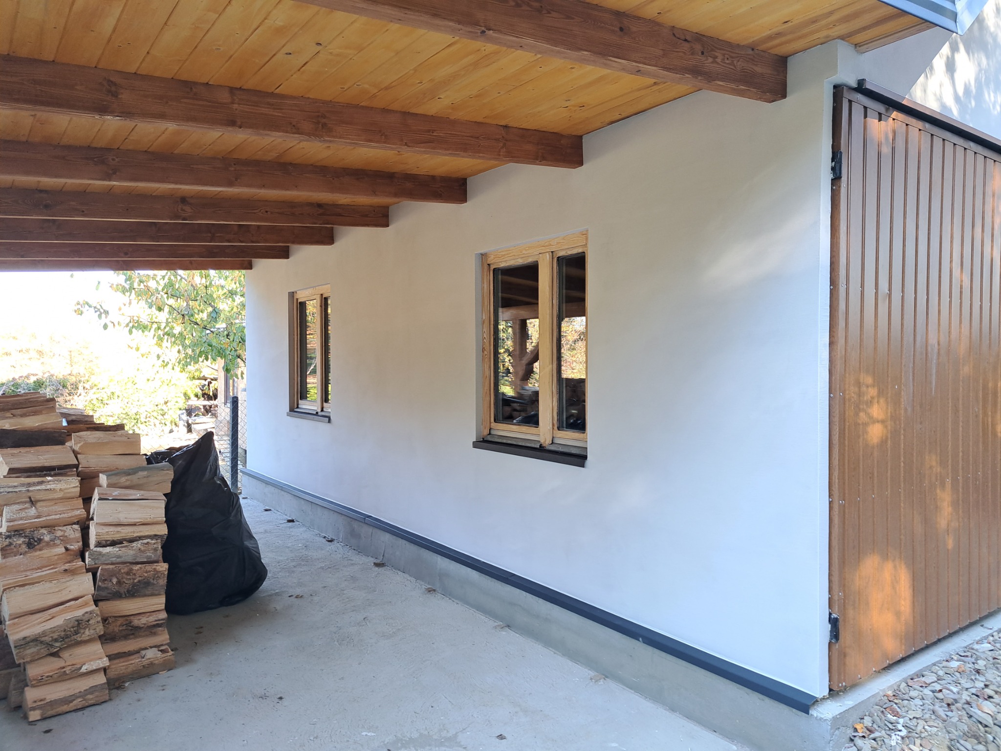 Wykończona elewacja budynku z drewnianymi oknami i przesuwną bramą garażową, widoczny stos drewna i czarny worek na zewnątrz.