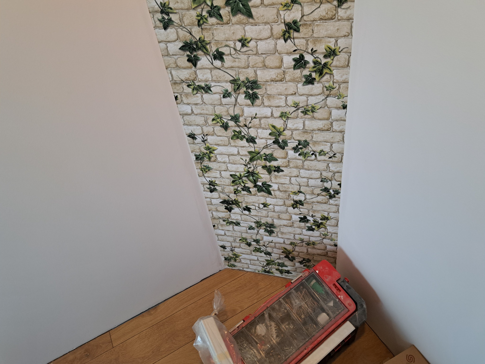 Fragment pokoju po położeniu tapety w cegiełki z motywem bluszczu na skośnej ścianie, widoczna skrzynka z narzędziami na podłodze.