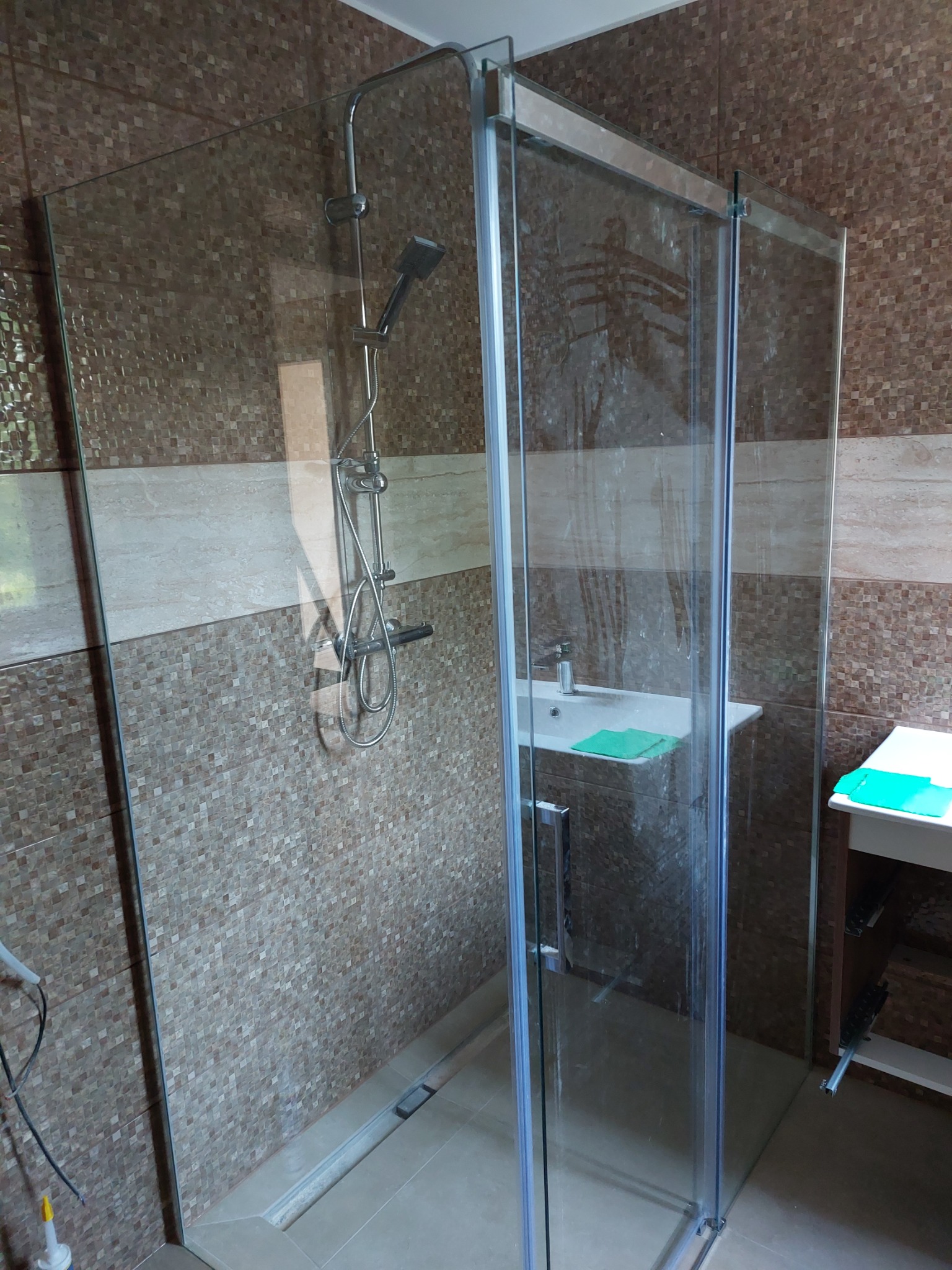 Szklana kabina prysznicowa z odpływem liniowym w nowocześnie wykończonej łazience, widoczna umywalka z zielonym ręcznikiem.