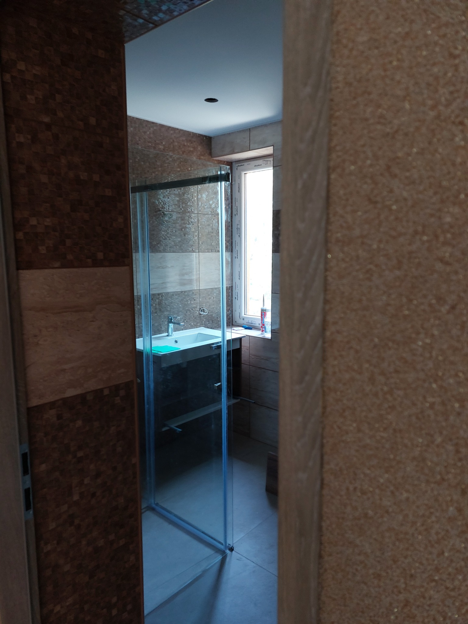 Nowoczesna łazienka z kabiną prysznicową ze szkła, umywalką z szafką i oknem, widok zza częściowo zasłoniętego wejścia.