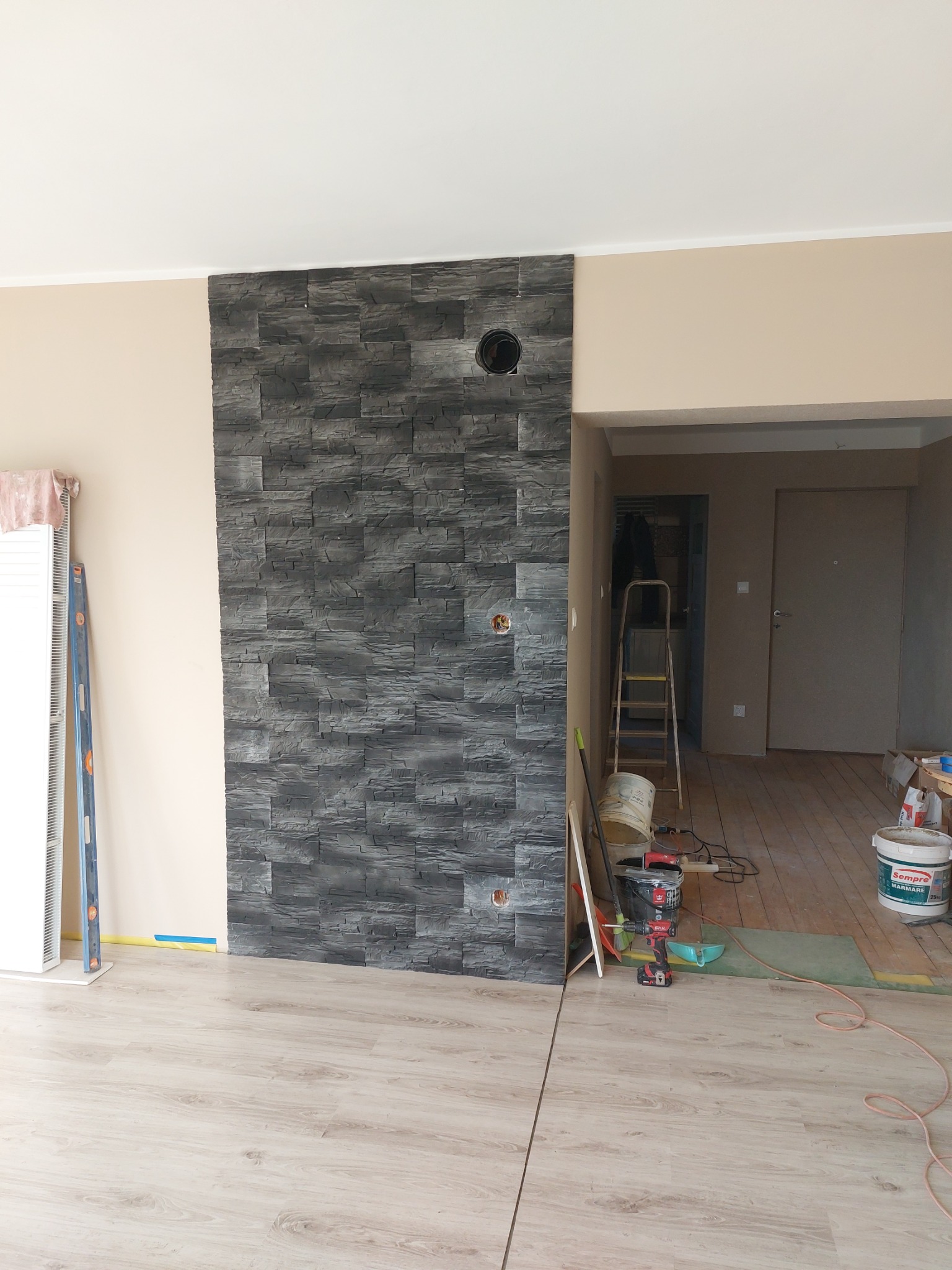 Fragment ściany z dekoracyjną okładziną kamienną w odcieniach szarości, z widocznymi otworami na gniazdka elektryczne, w trakcie prac wykończeniowych, z narzędziami i materiałami budowlanymi w tle.