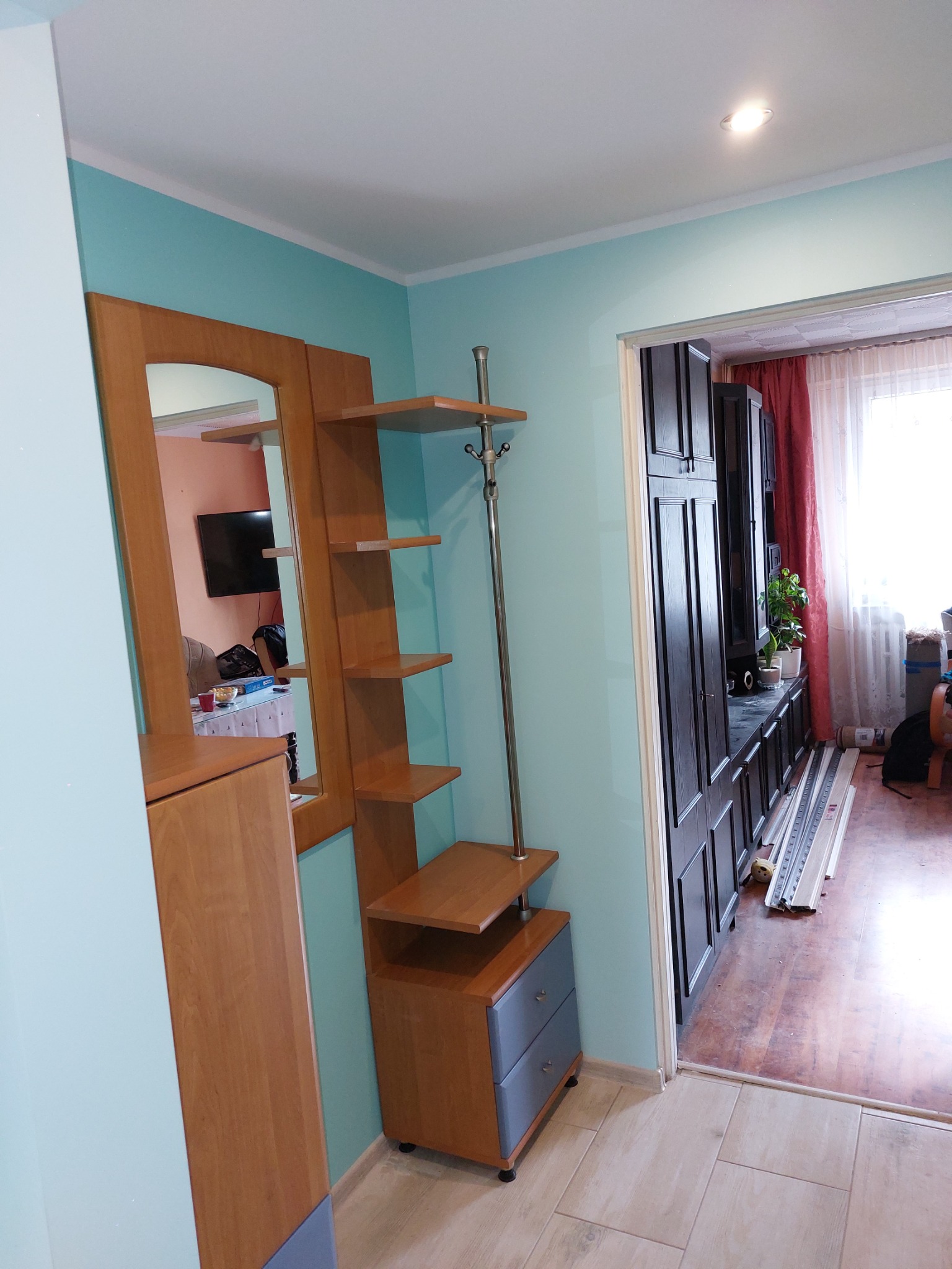 Przedpokój po remoncie, ściany pomalowane na turkusowo, drewniany zestaw mebli z lustrem i półkami, widoczna zabudowa meblowa w salonie, podłoga wyłożona jasnymi płytkami.