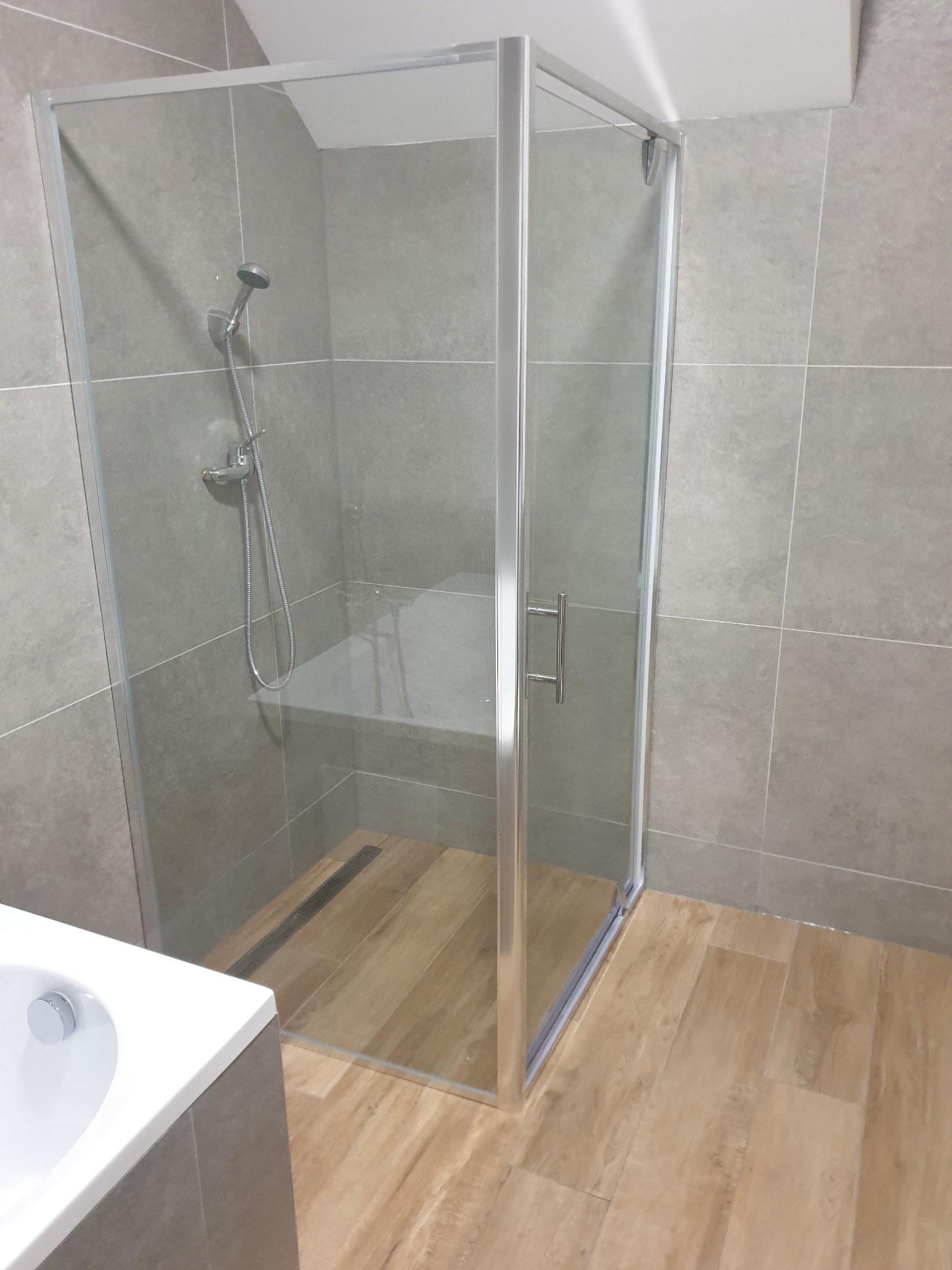 Szklana kabina prysznicowa z brodzikiem wyłożonym płytkami imitującymi drewno, zamontowana w łazience w Lesznie, wykończonej szarymi płytkami ściennymi.