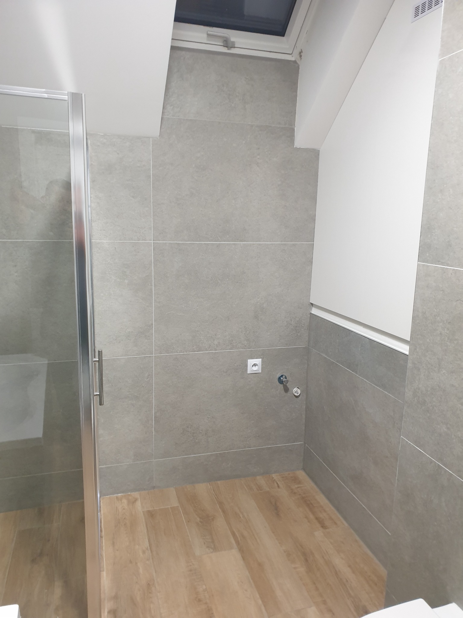 Wykończona łazienka na poddaszu z szarymi płytkami, drewnopodobną podłogą, kabiną prysznicową z chromowanymi okuciami i oknem dachowym.