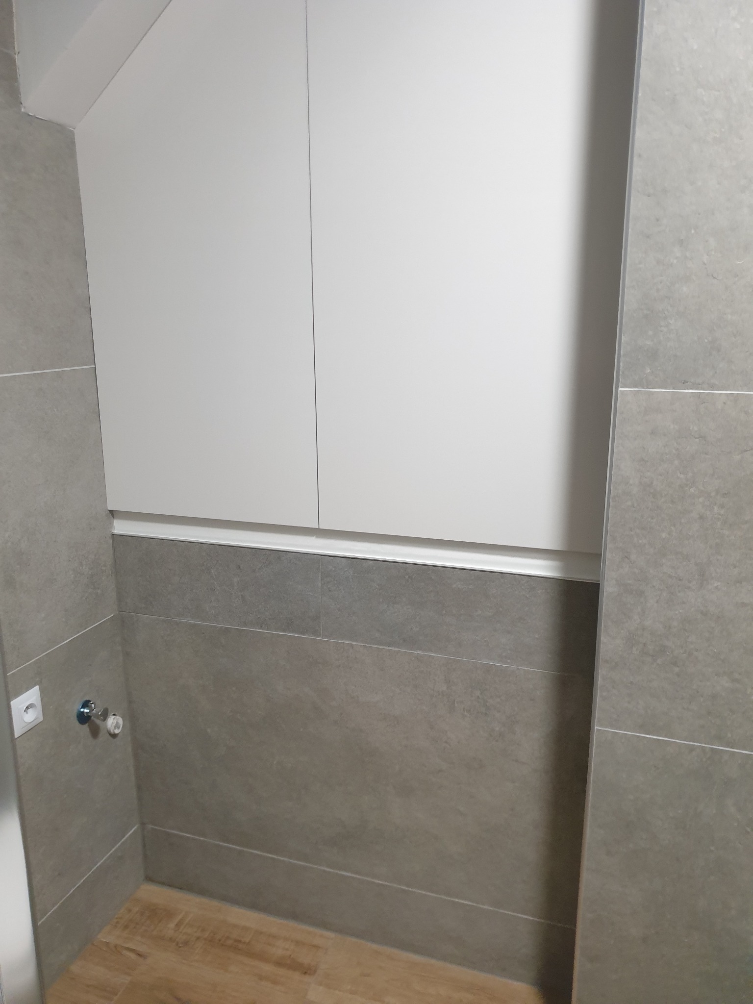 Wykończona łazienka z szarymi płytkami imitującymi kamień, drewnopodobną podłogą i białymi szafkami podwieszanymi pod skosem.