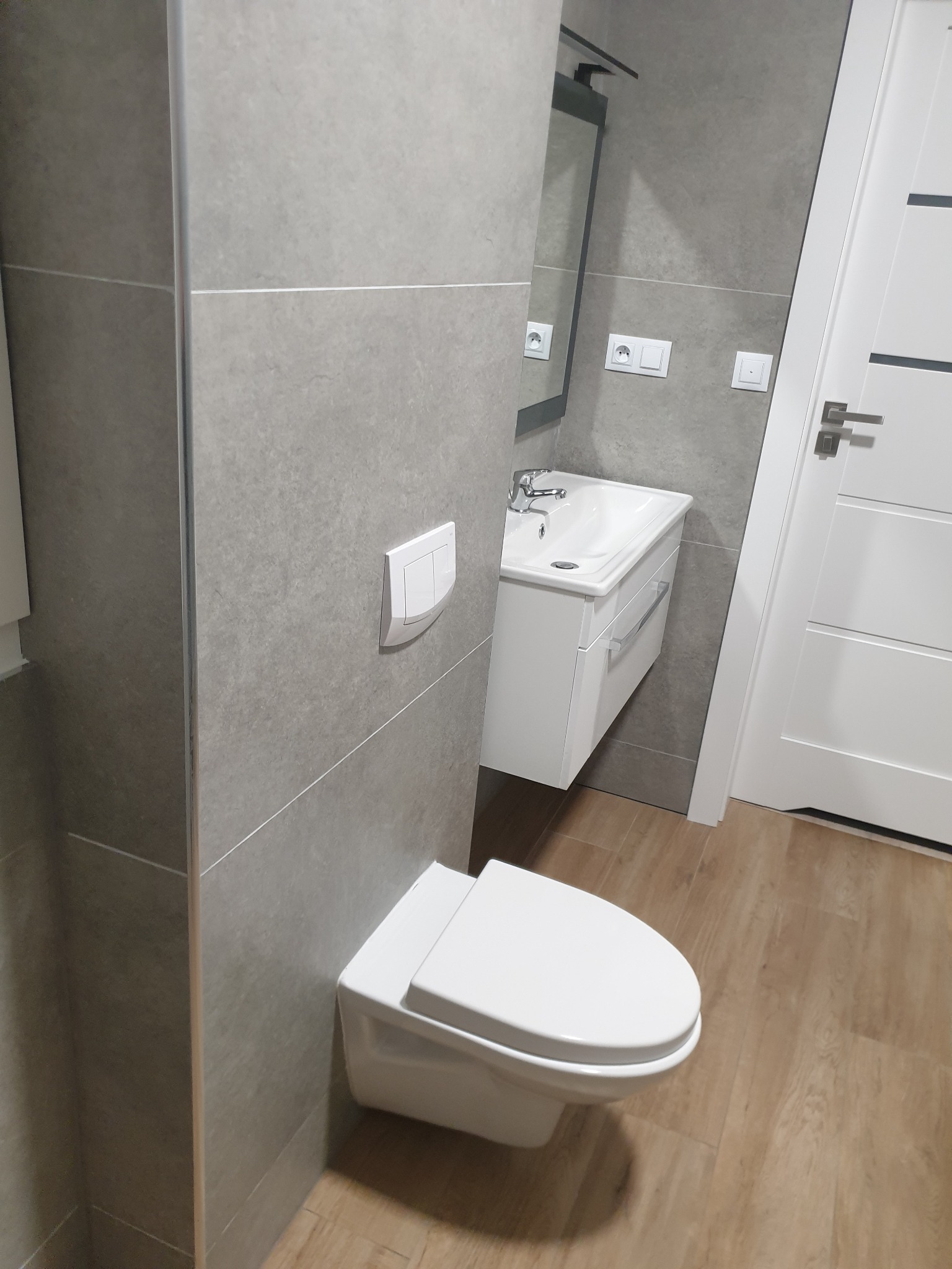 Nowoczesna łazienka z szarymi płytkami, podwieszaną toaletą, białą szafką z umywalką i drewnopodobną podłogą. Widoczny fragment lustra z oświetleniem.