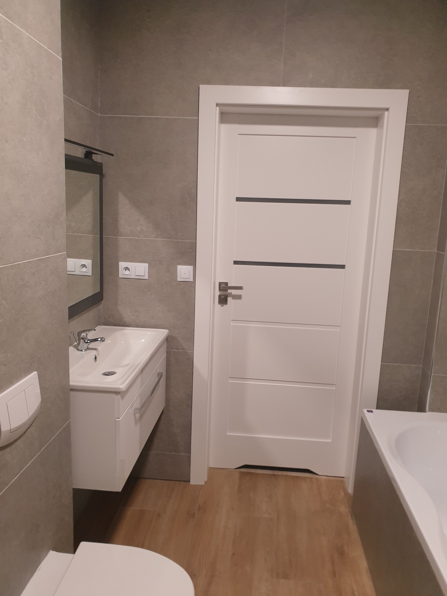 Wykończona łazienka z szarymi płytkami, białą umywalką z szafką, lustrem, toaletą i drzwiami z poziomymi paskami, widoczna również część wanny.