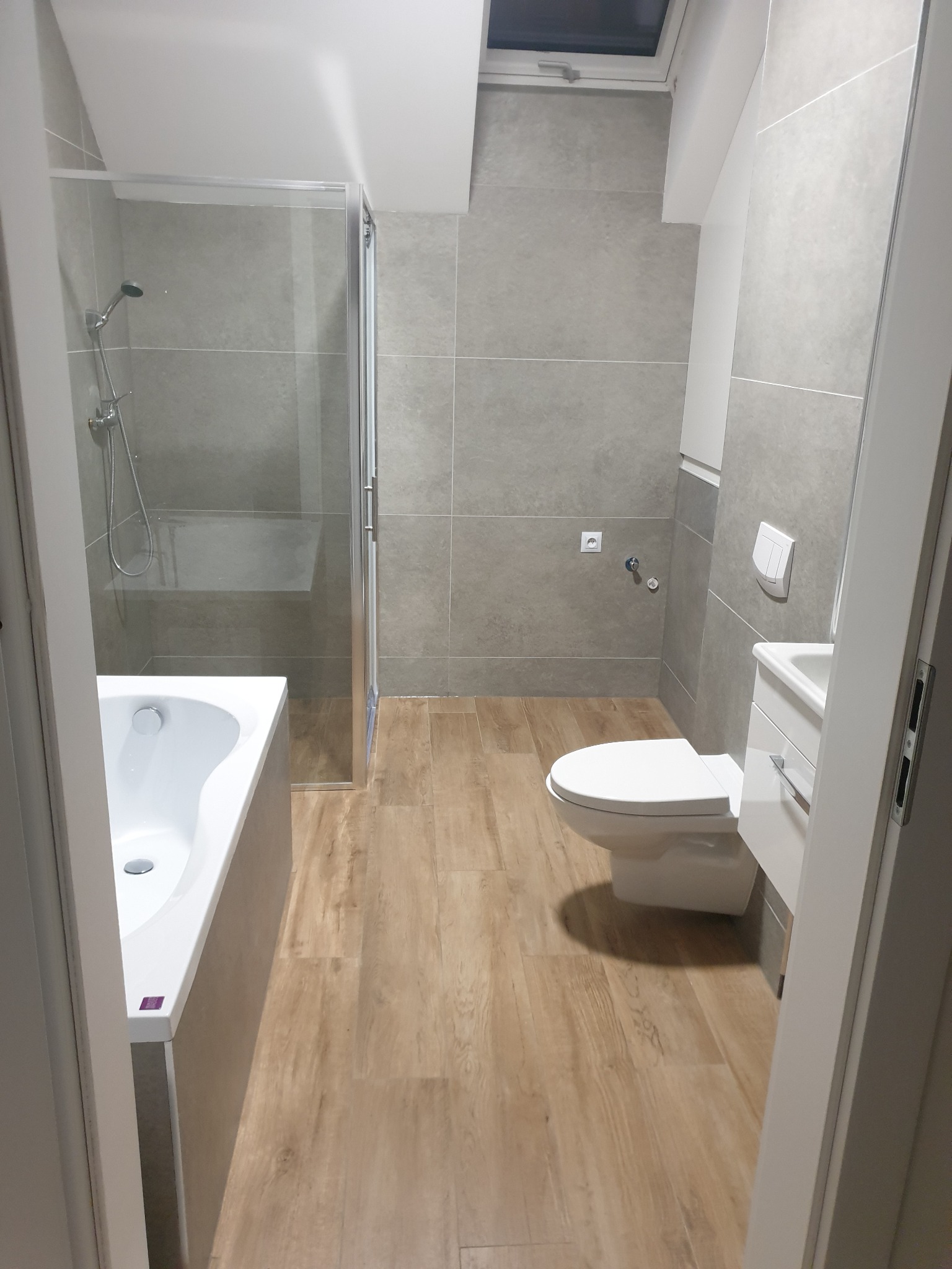 Nowoczesna łazienka z szarymi płytkami, drewnopodobną podłogą, białą wanną z parawanem, toaletą i umywalką z szafką, widziana zza futryny drzwi.