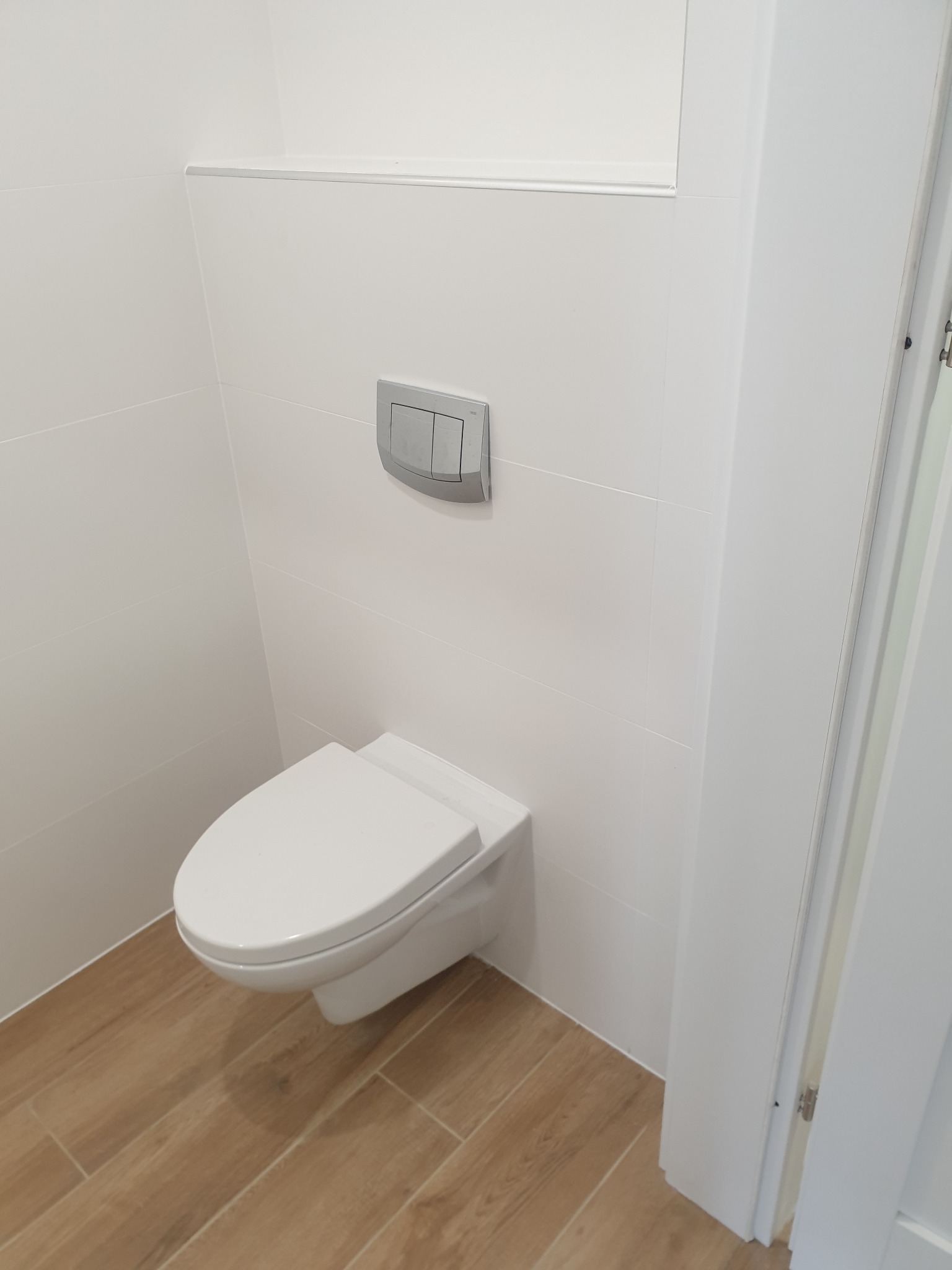 Nowoczesna toaleta podwieszana z chromowanym przyciskiem spłukującym, białe płytki ścienne i podłogowe imitujące drewno, widoczny fragment białych drzwi.