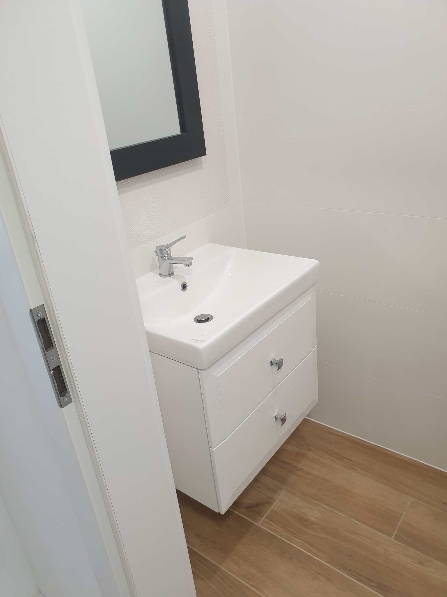 Nowoczesna łazienka z białą umywalką nablatową z szafką, chromowaną armaturą i lustrem w czarnej ramie, na podłodze płytki imitujące drewno.