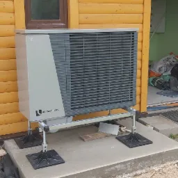 Szara pompa ciepła Alpha Innotec zamontowana na betonowym podeście przed drewnianym domkiem z widocznym wejściem i elementami instalacji wewnątrz.