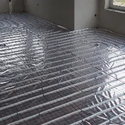 Białe rurki instalacji ogrzewania podłogowego ułożone spiralnie na srebrnej folii odblaskowej z czerwoną siatką, widoczne surowe ściany i fragment okna.