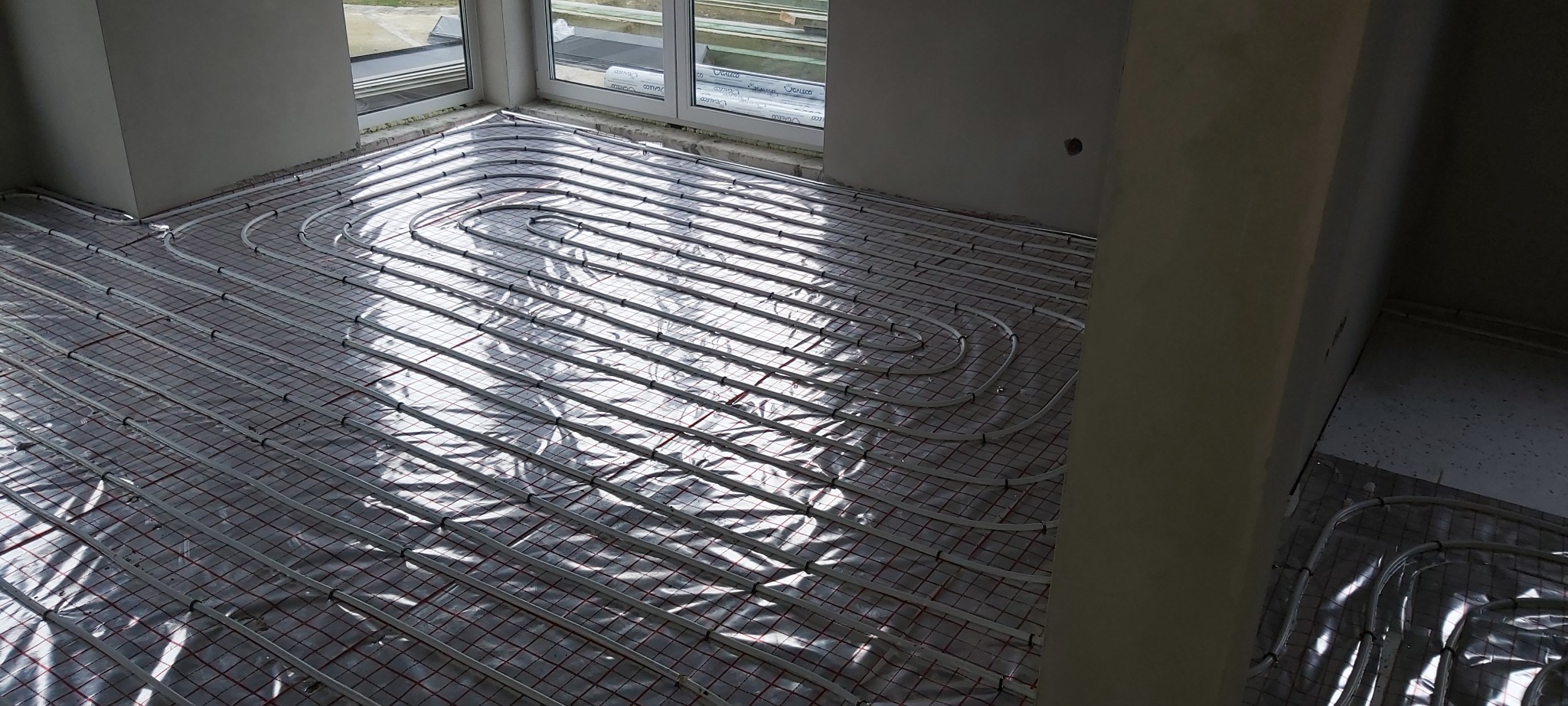 Rozłożone rury systemu ogrzewania podłogowego na folii izolacyjnej z siatką, widok z góry w surowym wnętrzu przed wylewką.