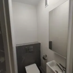 Nowoczesna toaleta z podwieszaną miską, szarą zabudową stelaża i umywalką w szafce, wykończona białymi płytkami imitującymi kamień, z widoczną wentylacją i lustrem.