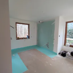 Wnętrze w trakcie prac: ściany częściowo pokryte płytami, zabezpieczona podłoga folią w kolorze turkusowym, widoczne instalacje elektryczne, okna z widokiem na las.