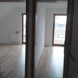 Widok przez dwa otwory drzwiowe na świeżo wykończone wnętrze w Złotowie: jasne ściany, drewniana podłoga, widoczne okna i grzejnik.