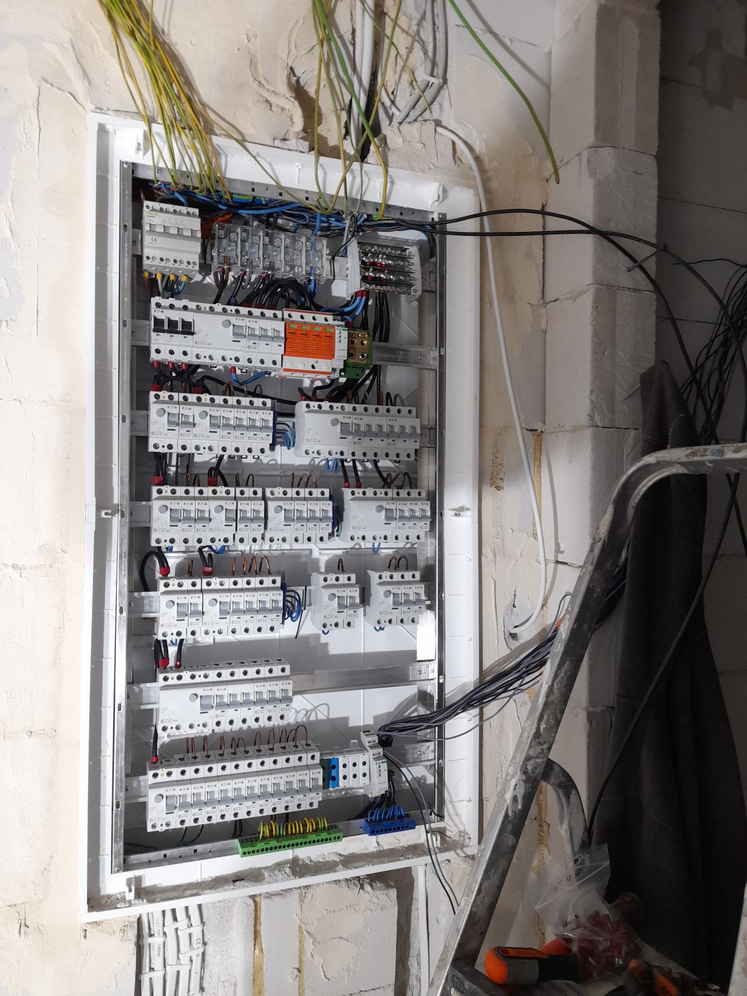 Rozdzielnica elektryczna z licznymi zabezpieczeniami i przewodami w surowym wnętrzu, widoczne ślady po kuciu w ścianie, drabina oparta o ścianę.